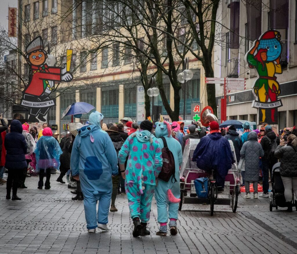 Straßenkarneval in Köln – Kostümierte in Tier-Overalls von hinten in einer Fußgängerzone, an den Häusern Figuren von Karnevalsgesellschaften