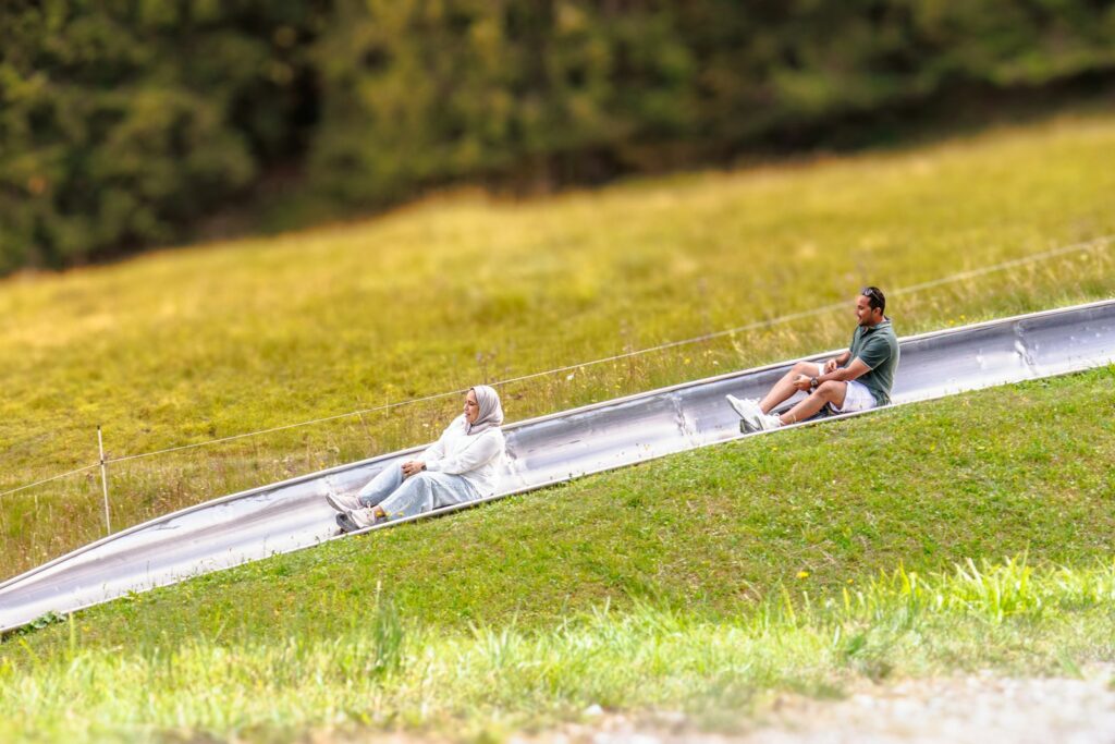 Zwei Personen fahren auf einer Sommerrodelbahn über eine grüne Wiese
